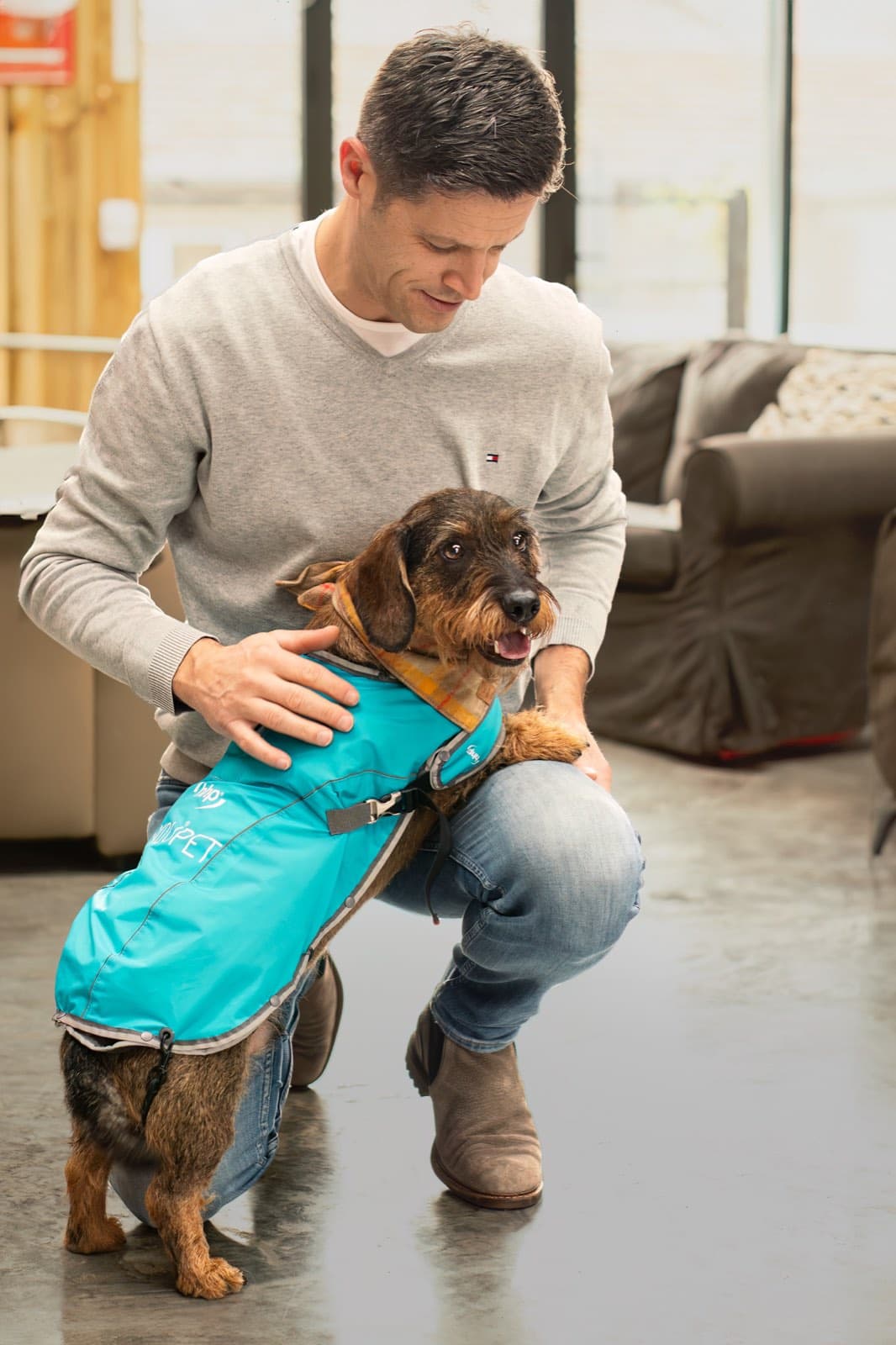 En husse som sitter på knä bredvid sin hund, som bär den turkosa ANDUPET-jackan.